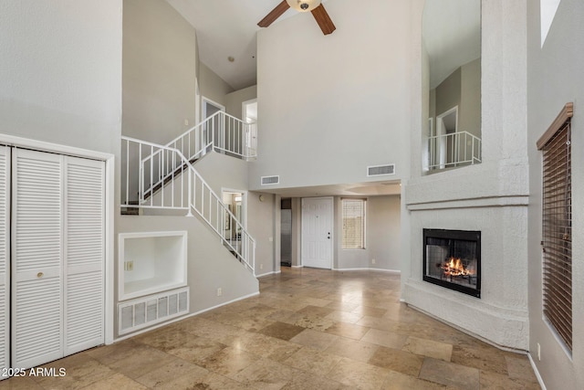 unfurnished living room featuring a towering ceiling, ceiling fan, a glass covered fireplace, stairway, and stone tile floors