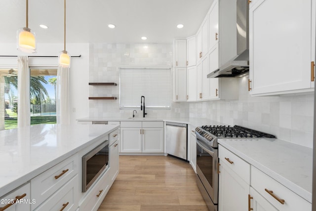 kitchen featuring wall chimney exhaust hood, sink, appliances with stainless steel finishes, pendant lighting, and white cabinets