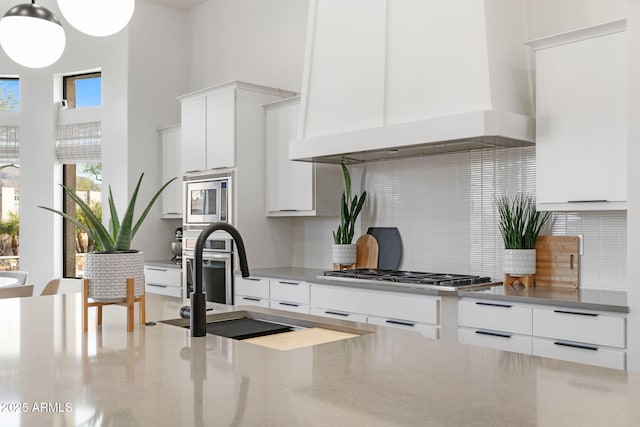 kitchen with a towering ceiling, white cabinets, backsplash, stainless steel appliances, and wall chimney range hood