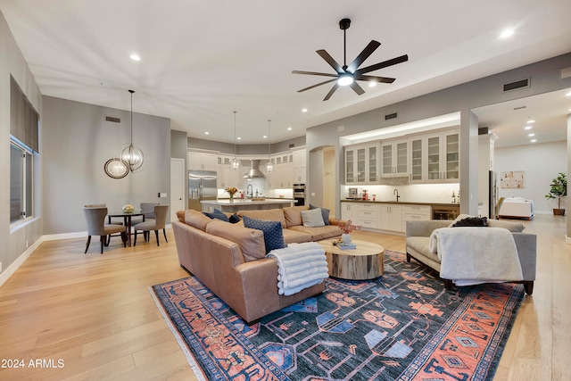 living room with wine cooler, ceiling fan with notable chandelier, sink, and light wood-type flooring