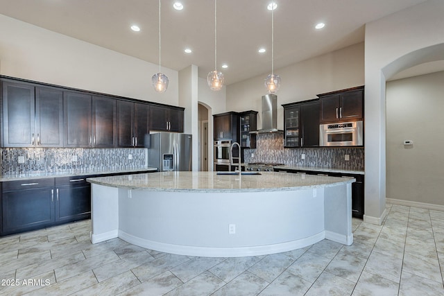 kitchen featuring arched walkways, a large island with sink, a sink, stainless steel appliances, and wall chimney range hood