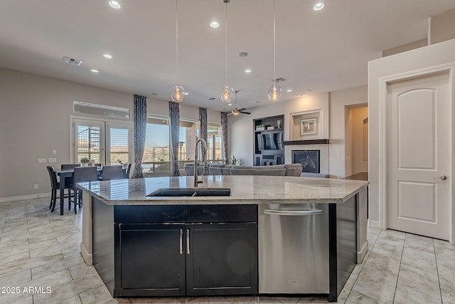 kitchen featuring light stone countertops, an island with sink, a fireplace, dark cabinetry, and a sink