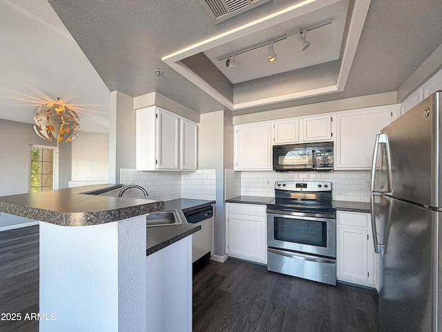 kitchen with a tray ceiling, stainless steel appliances, dark countertops, a sink, and a peninsula