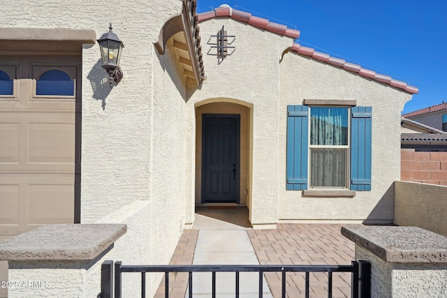 view of exterior entry with a garage, a tiled roof, fence, and stucco siding