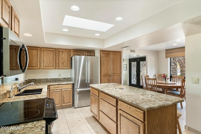 kitchen featuring a raised ceiling, appliances with stainless steel finishes, a sink, a kitchen island, and light stone countertops