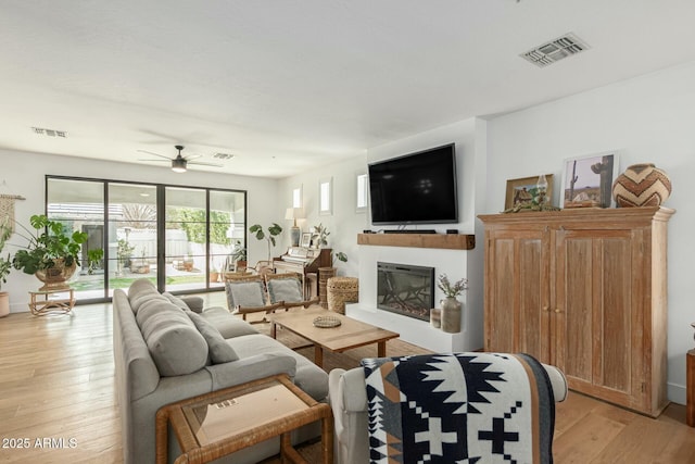 living area with light wood-type flooring, a glass covered fireplace, visible vents, and a ceiling fan