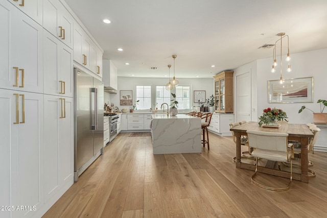 kitchen with light stone counters, white cabinetry, hanging light fixtures, high end appliances, and a center island with sink