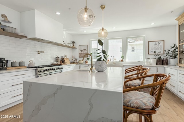 kitchen with light stone counters, double oven range, open shelves, an island with sink, and decorative light fixtures