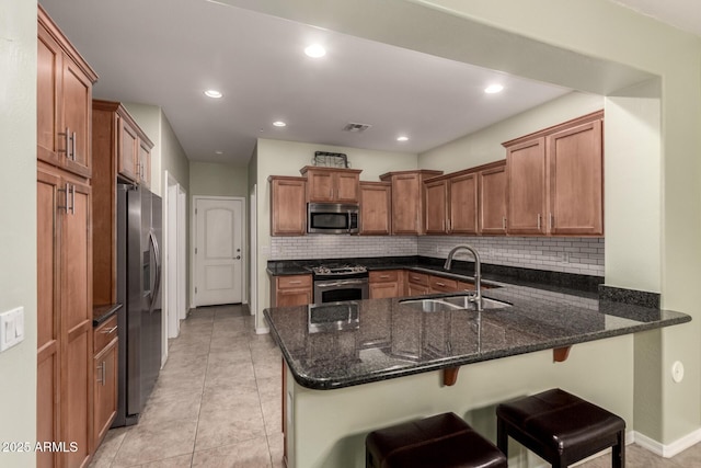 kitchen featuring a peninsula, stainless steel appliances, a sink, brown cabinets, and dark stone countertops