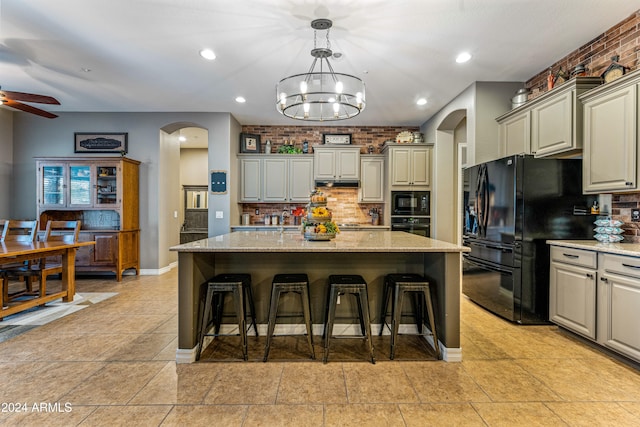 kitchen with light stone counters, gray cabinetry, ceiling fan with notable chandelier, a kitchen island with sink, and black appliances