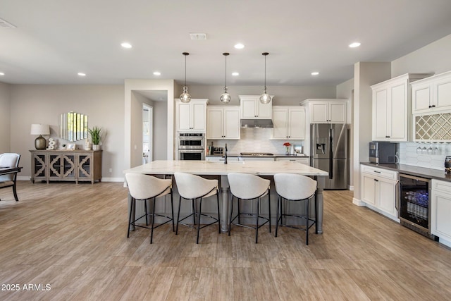 kitchen featuring appliances with stainless steel finishes, beverage cooler, an island with sink, and hanging light fixtures