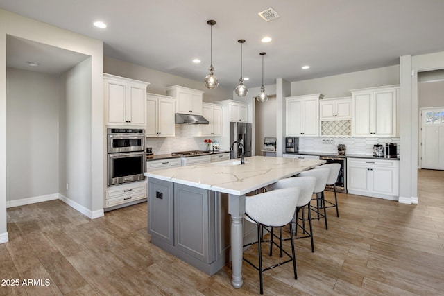 kitchen featuring stainless steel appliances, light stone countertops, an island with sink, white cabinets, and decorative light fixtures