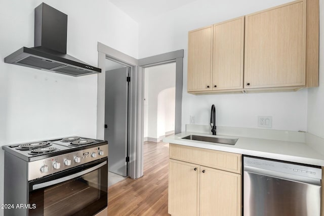 kitchen with light brown cabinetry, stainless steel appliances, sink, wall chimney range hood, and light hardwood / wood-style floors