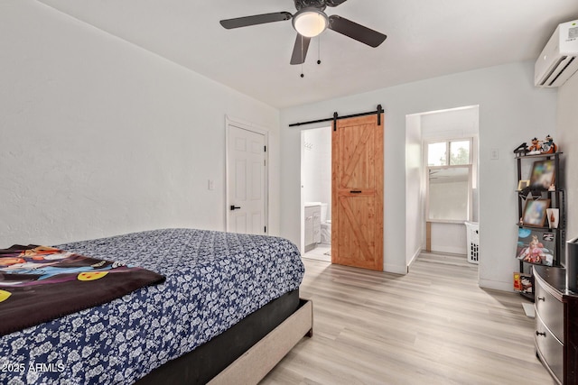 bedroom featuring connected bathroom, a wall mounted AC, light wood-type flooring, ceiling fan, and a barn door