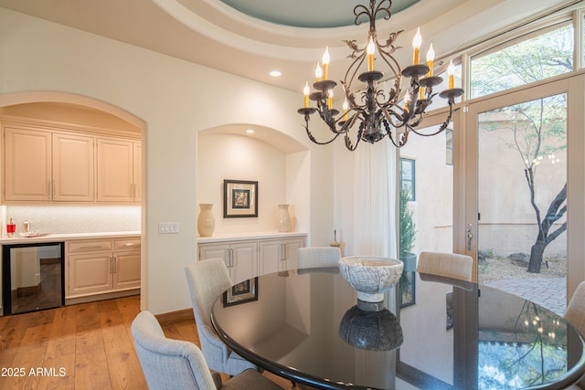 dining room featuring wine cooler, a notable chandelier, a tray ceiling, bar area, and light wood-type flooring