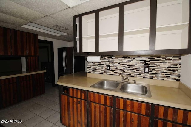 kitchen featuring sink, black fridge, light tile patterned floors, backsplash, and a drop ceiling