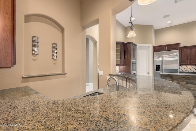 kitchen with appliances with stainless steel finishes, dark stone countertops, hanging light fixtures, dark brown cabinets, and a sink