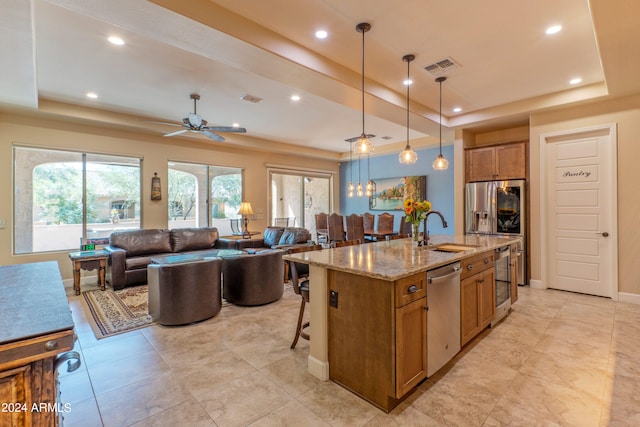 kitchen with a raised ceiling, a kitchen island with sink, stainless steel appliances, sink, and decorative light fixtures