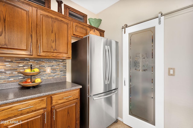 kitchen featuring dark countertops, backsplash, a barn door, freestanding refrigerator, and brown cabinetry