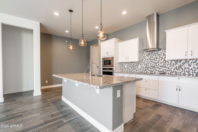 kitchen featuring wall chimney exhaust hood, stainless steel appliances, a kitchen island with sink, pendant lighting, and white cabinetry