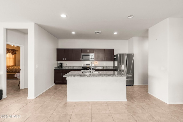 kitchen with appliances with stainless steel finishes, sink, a kitchen island with sink, light stone counters, and dark brown cabinetry