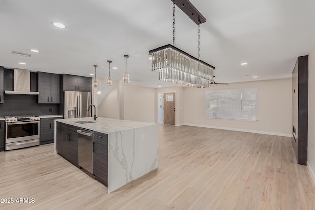 kitchen featuring wall chimney range hood, sink, appliances with stainless steel finishes, a kitchen island with sink, and hanging light fixtures