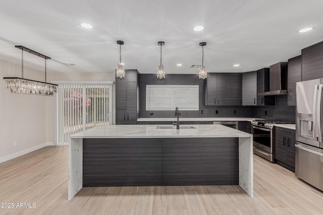 kitchen with sink, light stone counters, appliances with stainless steel finishes, a kitchen island with sink, and wall chimney range hood