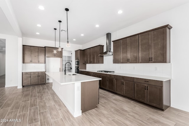 kitchen with dark brown cabinetry, wall chimney exhaust hood, hanging light fixtures, stainless steel gas stovetop, and a center island with sink