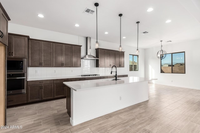 kitchen featuring sink, wall chimney exhaust hood, hanging light fixtures, stainless steel appliances, and a center island with sink