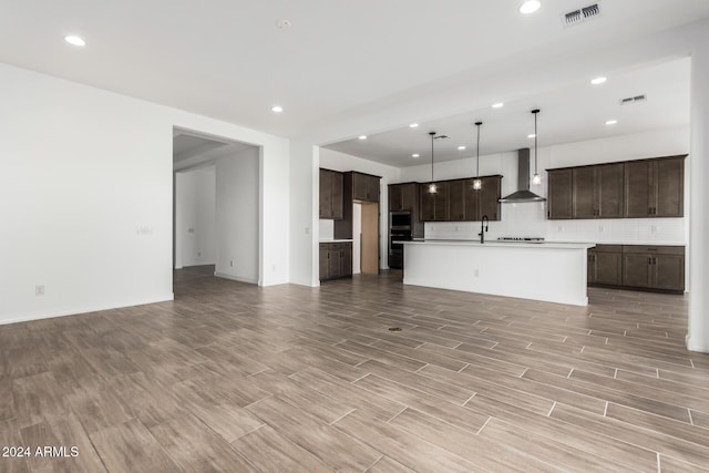 kitchen featuring pendant lighting, a kitchen island with sink, wall chimney range hood, decorative backsplash, and dark brown cabinets