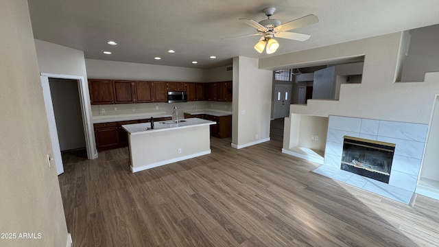 kitchen featuring a tile fireplace, a kitchen island with sink, wood-type flooring, ceiling fan, and sink