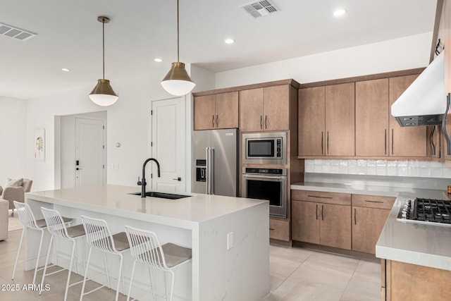 kitchen featuring sink, hanging light fixtures, appliances with stainless steel finishes, custom range hood, and a kitchen island with sink