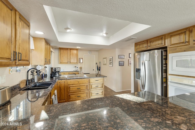 kitchen featuring kitchen peninsula, dark stone counters, white appliances, a tray ceiling, and sink