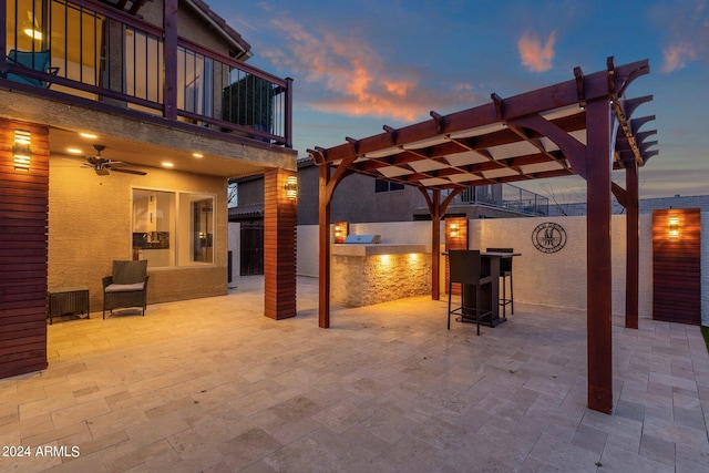 patio terrace at dusk with a bar, a pergola, ceiling fan, and a balcony