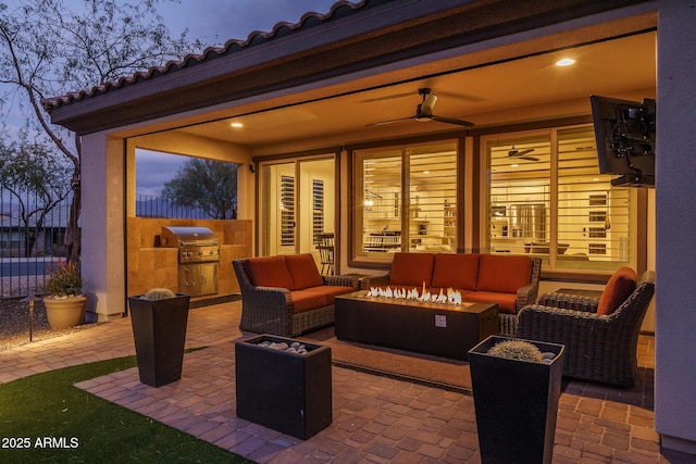 patio terrace at dusk featuring ceiling fan, an outdoor kitchen, an outdoor living space with a fire pit, and grilling area