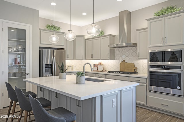 kitchen with pendant lighting, wall chimney range hood, sink, stainless steel appliances, and a center island with sink