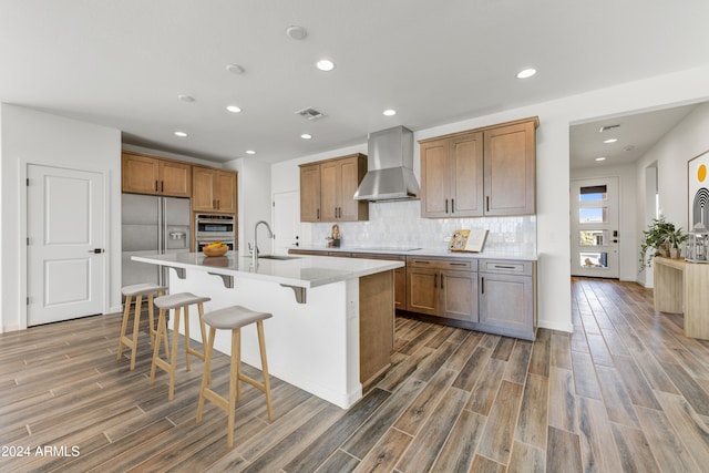 kitchen with wall chimney exhaust hood, dark hardwood / wood-style flooring, sink, stainless steel appliances, and an island with sink