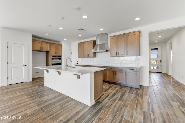 kitchen featuring wall chimney range hood, dark wood-type flooring, sink, an island with sink, and double oven