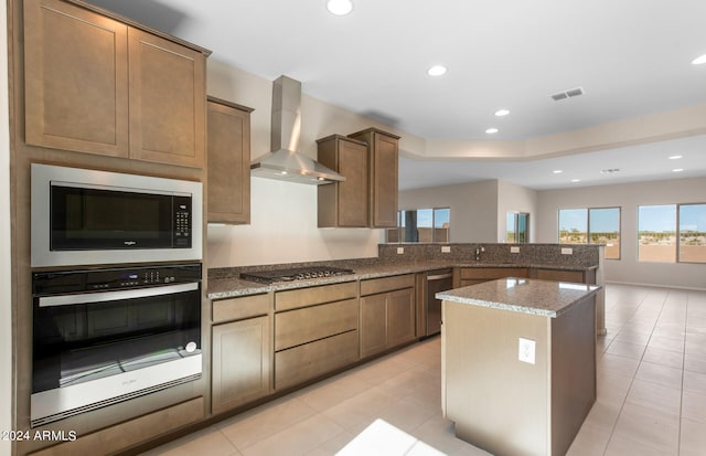 kitchen with stainless steel appliances, wall chimney range hood, dark stone counters, a center island, and light tile patterned flooring