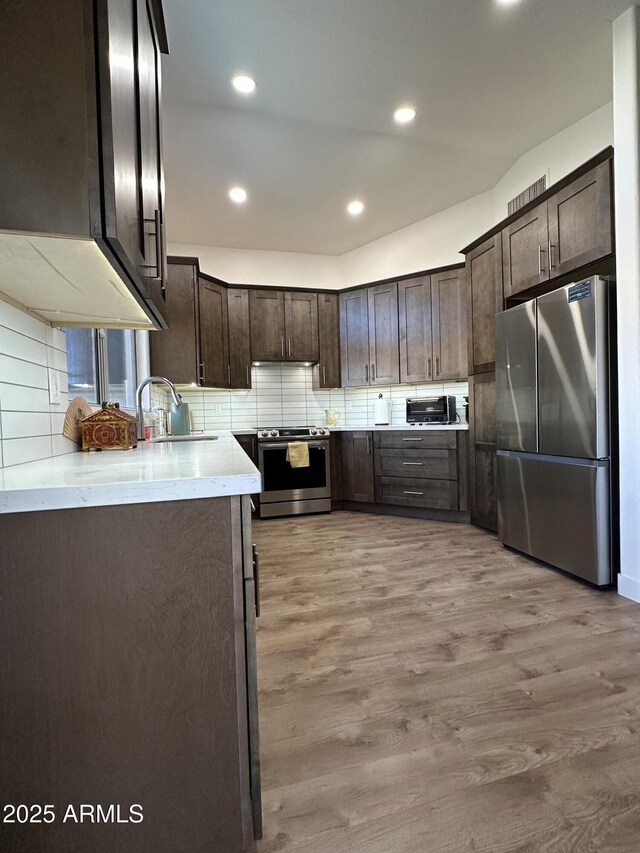 kitchen with light wood-style flooring, stainless steel appliances, a sink, dark brown cabinets, and decorative backsplash