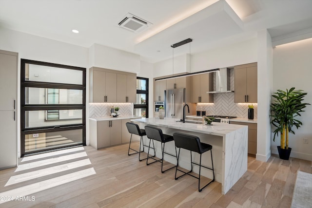 kitchen featuring sink, wall chimney exhaust hood, light wood-type flooring, an island with sink, and high end fridge