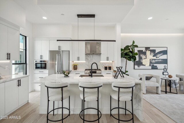 kitchen with a breakfast bar, stainless steel appliances, wall chimney range hood, pendant lighting, and a center island with sink