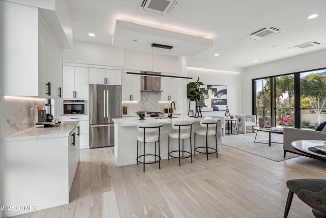 kitchen featuring stainless steel appliances, wall chimney range hood, tasteful backsplash, decorative light fixtures, and light wood-type flooring