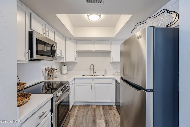 kitchen with light wood-type flooring, white cabinets, appliances with stainless steel finishes, a sink, and a raised ceiling