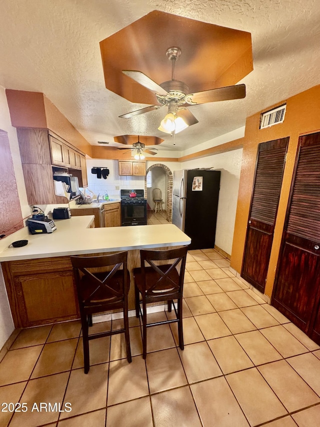 kitchen with light tile patterned flooring, black range with gas cooktop, a tray ceiling, stainless steel fridge, and kitchen peninsula