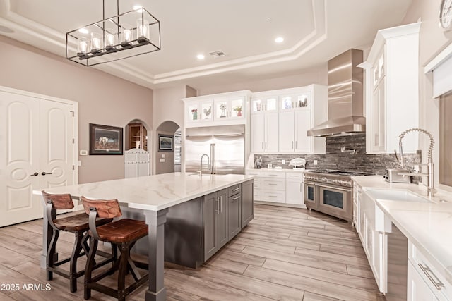 kitchen featuring high quality appliances, a raised ceiling, a center island with sink, and wall chimney range hood