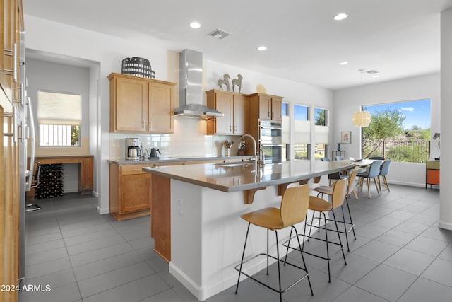 kitchen with tasteful backsplash, wall chimney range hood, a center island with sink, sink, and light tile patterned floors