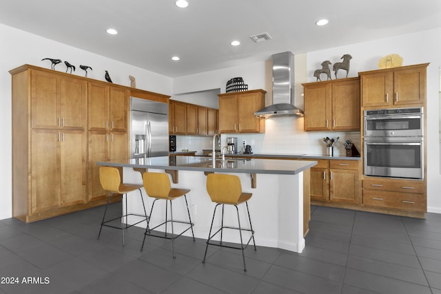 kitchen featuring appliances with stainless steel finishes, wall chimney exhaust hood, sink, a kitchen breakfast bar, and a center island with sink