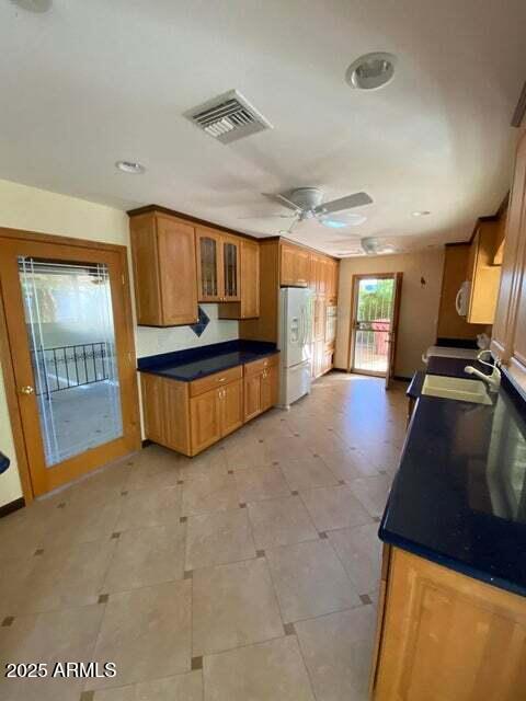 kitchen with freestanding refrigerator, brown cabinets, a ceiling fan, glass insert cabinets, and dark countertops