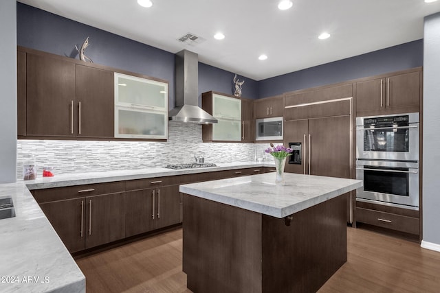 kitchen featuring tasteful backsplash, a kitchen island, dark wood-type flooring, wall chimney range hood, and appliances with stainless steel finishes